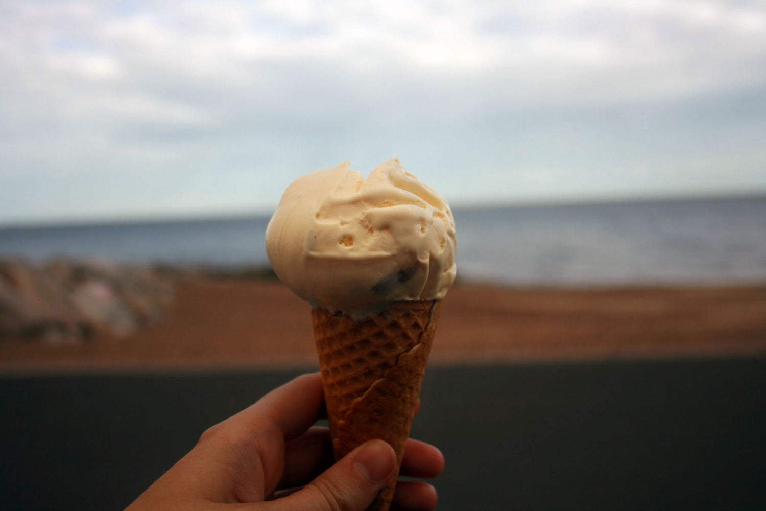 Someone holding a vanilla ice cream in the foreground, and in the background a beach.